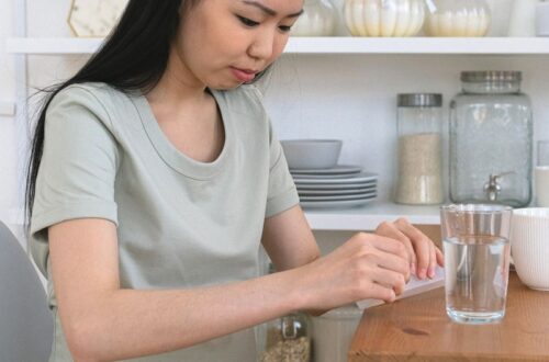 Pregnant woman in a sage green dress at a wooden kitchen table pressing a tablet out of a blister pack beside a glass of water