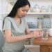 Pregnant woman in a sage green dress at a wooden kitchen table pressing a tablet out of a blister pack beside a glass of water