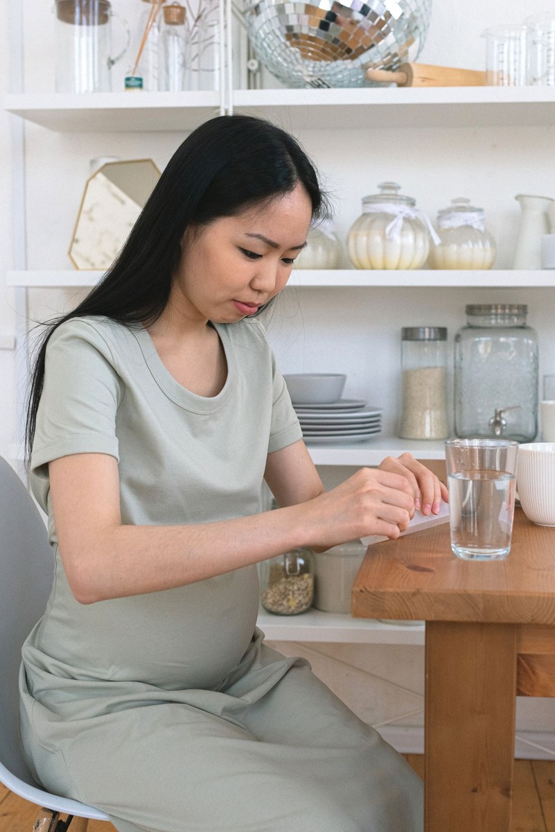 Pregnant woman in a sage green dress at a wooden kitchen table pressing a tablet out of a blister pack beside a glass of water